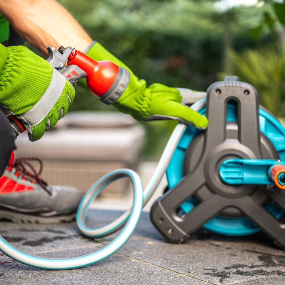 A person wearing gloves connects a garden hose from a reel to water plants in a sunny backyard.