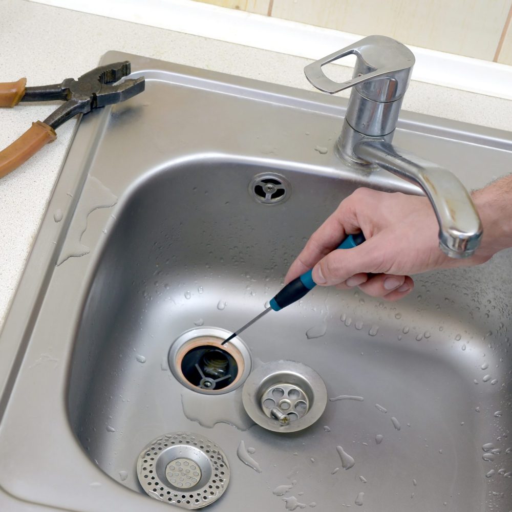 Close-up of handsome plumber repairing drain of kitchen sink. Photo of metalic sink and handyman with screwdriver